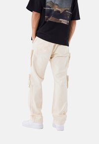 Pantalones cargo beige con bolsillos laterales, un corte relajado y tejido duradero; combinados con zapatillas deportivas blancas y una camiseta gráfica negra.