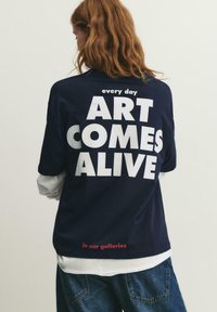 Person mit langen Haaren trägt ein dunkelblaues Hemd mit großem weißem Text „ART COMES ALIVE“ und kleineren Texten „every day“ und „in our galleries.“