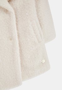 Weißer Mantel mit Struktur aus weichem Stoff, ausgestattet mit einem Reverskragen, einer Brusttasche und einem kleinen metallischen Logo-Akzent am Ärmel.