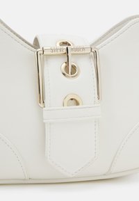 Ceinture en cuir blanc avec une boucle en métal doré gravée "Steve Madden" et des œillets dorés sur un fond lisse.