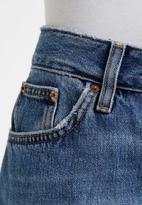 Džínové kalhoty s vysokým pasem, s roztrhaným lemem, měděnými nýty a zlatým šitím. Světle modrá barva s texturovaným povrchem.