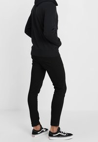 Sudadera negra con capucha y bolsillo delantero, combinada con jeans negros ajustados y zapatillas Vans negras. El atuendo presenta un diseño casual.