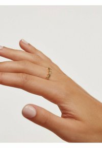 Bague en or au design élancé, ornée de multiples petites pierres de couleur claire disposées en courbe, portée sur une main avec un vernis à ongles neutre.