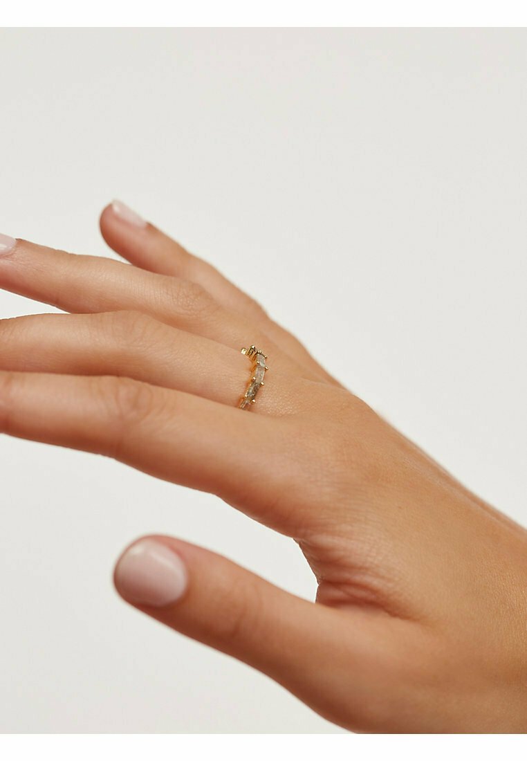 Bague en or au design élancé, ornée de multiples petites pierres de couleur claire disposées en courbe, portée sur une main avec un vernis à ongles neutre.