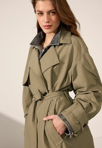 Trench coat color khaki con vita aderente, colletto oversize e rivestimento grigio. Presenta grandi tasche frontali e dettagli con bottoni sulle maniche.