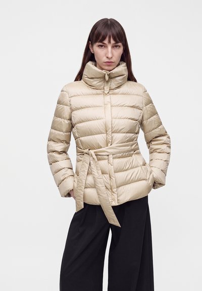 Femme portant une doudoune beige matelassée avec un col montant et une ceinture, associée à un pantalon noir ample, debout les mains dans les poches.