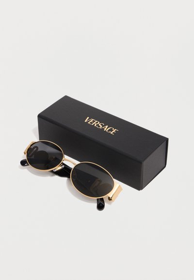 Οβάλ γυαλιά ηλίου με μαύρους φακούς και χρυσά πλαίσια, ακουμπισμένα δίπλα σε μαύρο κουτί με ανάγλυφο το χρυσό λογότυπο "VERSACE" σε λευκό φόντο.