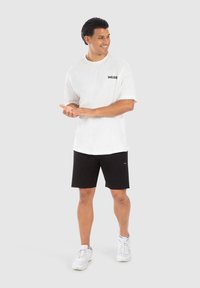 Hvid t-shirt med korte ærmer og "SMILODO" logo, parret med sorte shorts og hvide sneakers. Glat stof, afslappet pasform, casual design.