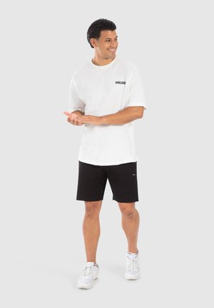 Weißes Kurzarm-T-Shirt mit "SMILODO"-Logo, kombiniert mit schwarzen Shorts und weißen Sneakers. Glatter Stoff, lockere Passform, lässiges Design.