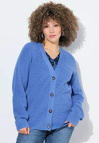 Modrý kardigan s volným střihem, žebrovanou texturou a šesti velkými černými knoflíky, vrstvený přes černý vzorovaný top, v kombinaci s džíny.