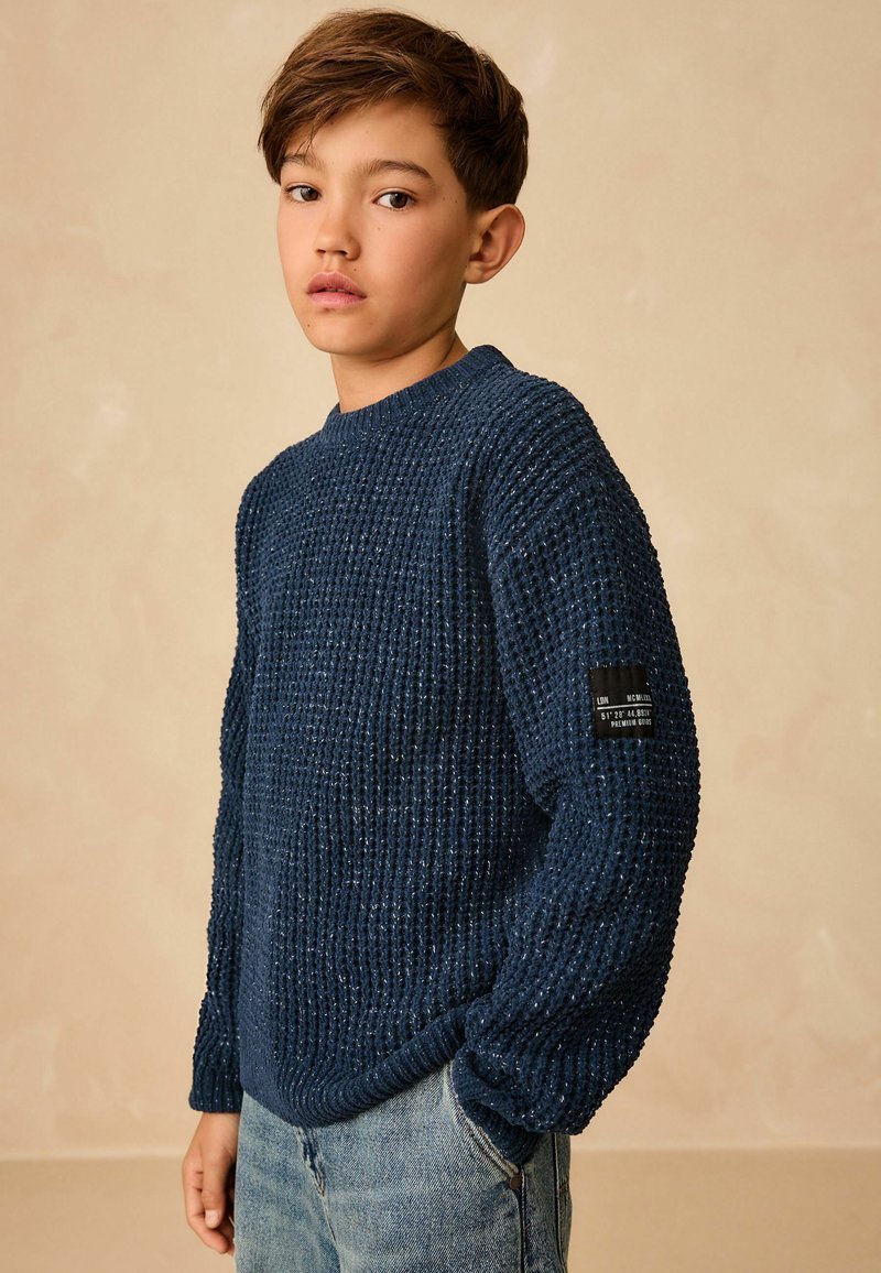 Maglione a maglia blu navy con superficie texturizzata, scollatura rotonda, polsini a coste e una toppa con logo sulla manica, abbinato a jeans azzurri chiari.