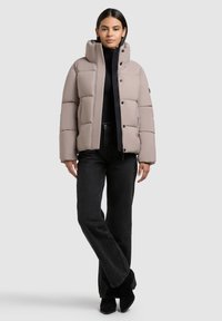 Beige puffjakke med høj krave, med vandret quiltede sektioner og en sort lynlås. Sammen med mørke jeans med høj talje.