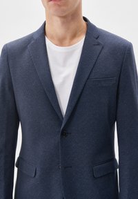 Navy blazer made of textured fabric, featuring a single-button closure, notch lapels, and flap pockets. Worn over a white t-shirt.