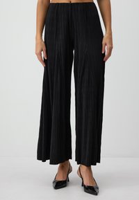 Pantalons larges plissés noirs avec une taille élastique, présentant des lignes verticales et une texture lisse, associés à des talons aiguilles noirs.
