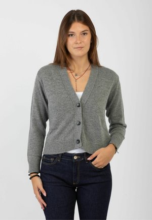 Giovane donna che indossa un cardigan grigio con bottoni sopra una maglietta bianca e jeans blu scuro, in piedi con una mano in tasca contro uno sfondo semplice.