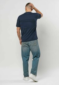 Mann i marineblå t-skjorte, blå jeans og hvite joggesko står med ryggen til kameraet, hånd berører bakhodet.