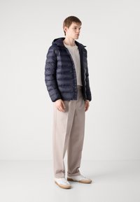 Giacca puffer blu navy con cappuccio, design trapuntato, chiusura frontale con zip, indossata sopra una maglietta grigia chiara, abbinata a pantaloni beige e sneakers bianche.