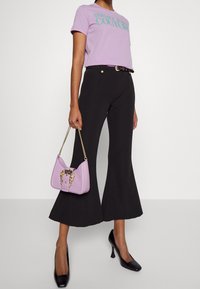 Kvinna som bär en lavendelfärgad Versace Couture t-shirt, svarta utsvängda byxor, svarta klackar och håller en lavendelfärgad handväska med guldkedjesnodd.