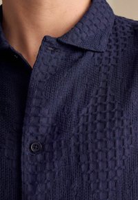 Camisa azul marino con un tejido texturizado con un patrón de cuadrícula, cuello puntiagudo y un detalle de botón negro.