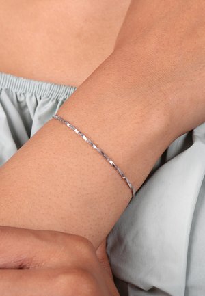 Bracelet en argent avec un design texturé et géométrique. Le bracelet est fin, métallique et repose sur un bras bronzé sous un tissu doux.