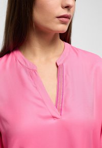 Blusa rosa con scollatura a V caratterizzata da un sottile dettaglio a righe. Tessuto liscio e lucido con una morbida consistenza, maniche corte e cuciture minime.