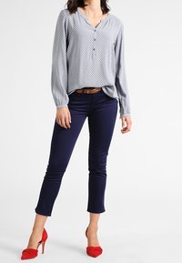 Vaaleansininen kuviollinen paita, jossa on lovikaulus ja nappisulku; yhdistettynä tummansinisiin istuviin housuihin ja punaisiin korkokenkiin.