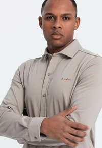 Beige poloko s dlouhým rukávem vyrobené z měkké tkaniny, s límcem na knoflíčky, předními knoflíky a jemně vyšitým logem na hrudi.