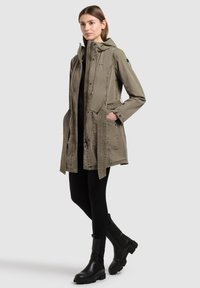 Femme portant un manteau à capuche vert olive avec ceinture, des leggings noirs et des bottines noires, debout devant un fond uni.