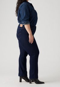 Mörka denimjeans med rak passform, hög midja och ikonisk bakfickdesign. Bärs med en blå denimskjorta och svarta klackstövlar.
