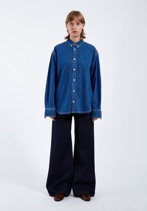 Jeanshemd in Mittelblau mit Knopfleiste, Kragen und langen Ärmeln. Kombiniert mit weit geschnittenen, dunklen Jeans. Glatte Textur, lässige Passform.