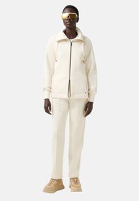 Crèmekleurige zip-up jas met een hoge kraag, gecombineerd met bijpassende broek. Accenten zijn onder meer structuurmateriaal en beige chunky sneakers.