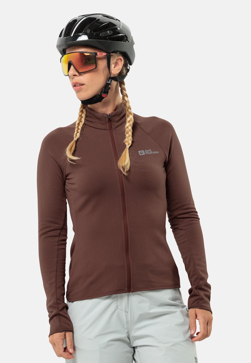 Giacca marrone con zip in un tessuto elasticizzato, con colletto alto e maniche lunghe, abbinata a pantaloni grigi chiari. Presenta un logo riflettente.