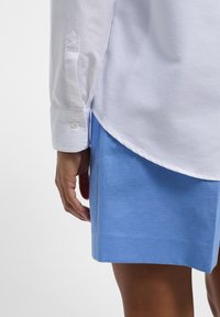 Camicia bianca a maniche lunghe con polsini abbottonati, abbinata a una gonna di lino blu chiaro. Texture morbida, orlo curvo e dettagli di design casual visibili.