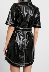 Svart, glansig kort klänning med korta ärmar, en krage och ett matchande bälte. Har synlig sömnad längs sömmarna.