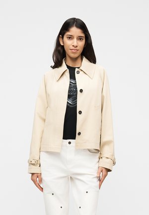 Kvinde med langt mørkt hår iført en beige jakke med knapper over en sort grafisk skjorte og hvide bukser, stående mod en ensfarvet baggrund.