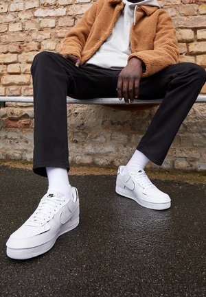 Baskets blanches Nike Air Force 1 avec embout perforé, accents noirs, tige en cuir texturé et semelle en caoutchouc blanc, portées avec un pantalon noir et des chaussettes noires.