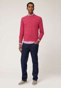 Maglione rosso lavorato a maglia con un motivo a rombi, indossato sopra una camicia a righe bianca e rosa, abbinato a jeans denim scuri e scarpe grigie.