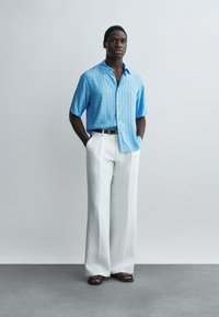 Άνδρας στέκεται με τα χέρια στις τσέπες, φορώντας ανοιχτό μπλε πουκάμισο με σχέδια και κοντά μανίκια, λευκό παντελόνι με φαρδιά πόδια, μαύρη ζώνη και σκούρα σανδάλια σε γκρι δάπεδο.