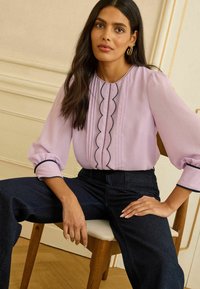 Vaalea laventelinvärinen pusero, jossa on pussihihat, mustat aaltoilevat yksityiskohdat ja nappikoristeet. Yhdistetty tummiin denimhousuihin.