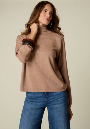 Kvinde iført en glitrende beige turtleneck sweater, blå jeans, store hoop-øreringe og chunky armbånd på beige baggrund.