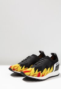 Zapatillas con una parte superior sintética negra, patrón de llamas amarillas y rojas en la punta, suela de goma texturizada y logotipo de marca en blanco en el costado.
