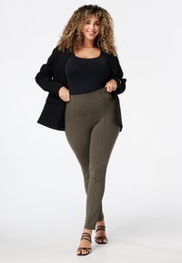 Femme aux cheveux bouclés portant un blazer noir, un haut noir, un pantalon taille haute olive avec des boutons et des talons noirs à lanières, se tenant avec confiance.