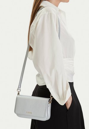Frau trägt weiße Bluse und schwarze Hose, trägt eine hellgraue Umhängetasche mit goldenen Details auf der Schulter.