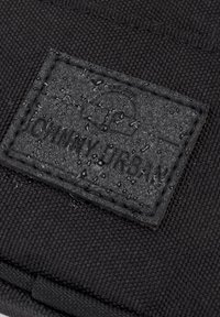 Detailní záběr na černou texturovanou tkaninu s našitým obdélníkovým záplatou pokrytým kapkami vody, na které je vyražený text značky.