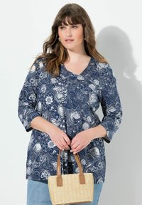 Frau in blauem blumigen Blusenshirt und hellblauen Jeans, die eine kleine, geflochtene Handtasche mit braunen Lederriemen hält.