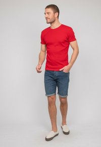 T-shirt rouge à manches courtes associé à un short en denim bleu, présentant des poignets roulés et des chaussures décontractées à enfiler avec une tige en toile de couleur claire.