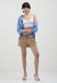 Camicia button-up azzurro chiaro in pizzo, canotta bianca corta, pantaloni corti color beige e sandali bianchi con cinturini incrociati. Bracciali metallici al polso.