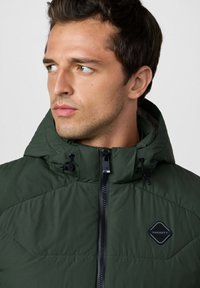 Giacca isolante verde scuro con cappuccio, dotata di zip frontale, lacci elastici e una patch in gomma nera. Tessuto testurizzato.