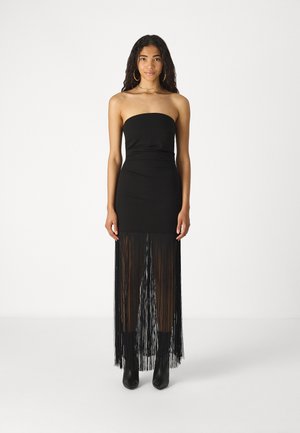 Black strapless dress with a fitted bodice, flowing fringe detail at the hem, and sleek texture. Worn with black pointed ankle boots.