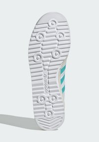 Biela podrážka tenisky s cirkulárnymi trakčnými bodmi, horizontálnymi drážkami a čiastočne viditeľným horným dielom so zelenými pruhmi na svetlom pozadí.